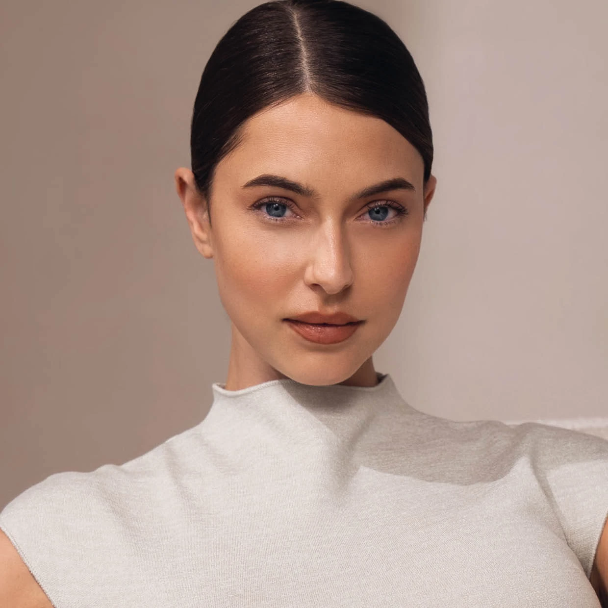 Porträt eines Models mit sleek bun, natürlichem Make-up und klarem Blick in die Kamera vor einem warmen, minimalistischen Studiohintergrund.