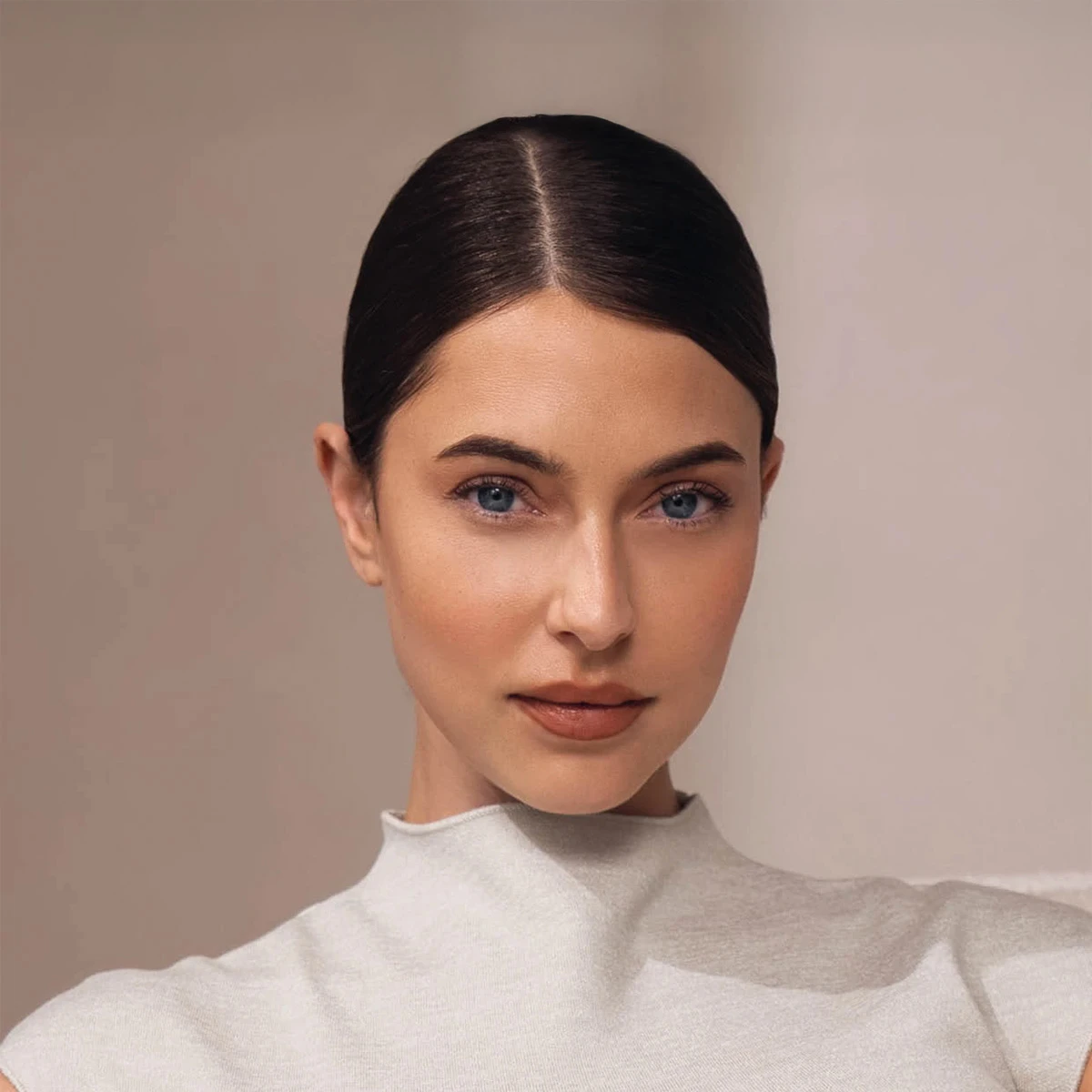 Porträt eines Models mit sleek bun, natürlichem Make-up und klarem Blick in die Kamera vor einem warmen, minimalistischen Studiohintergrund.